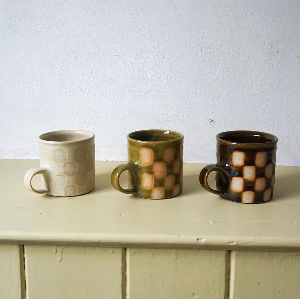 Three Edmund Davies checkerboard mugs in cream, green and brown glazes on a shelf