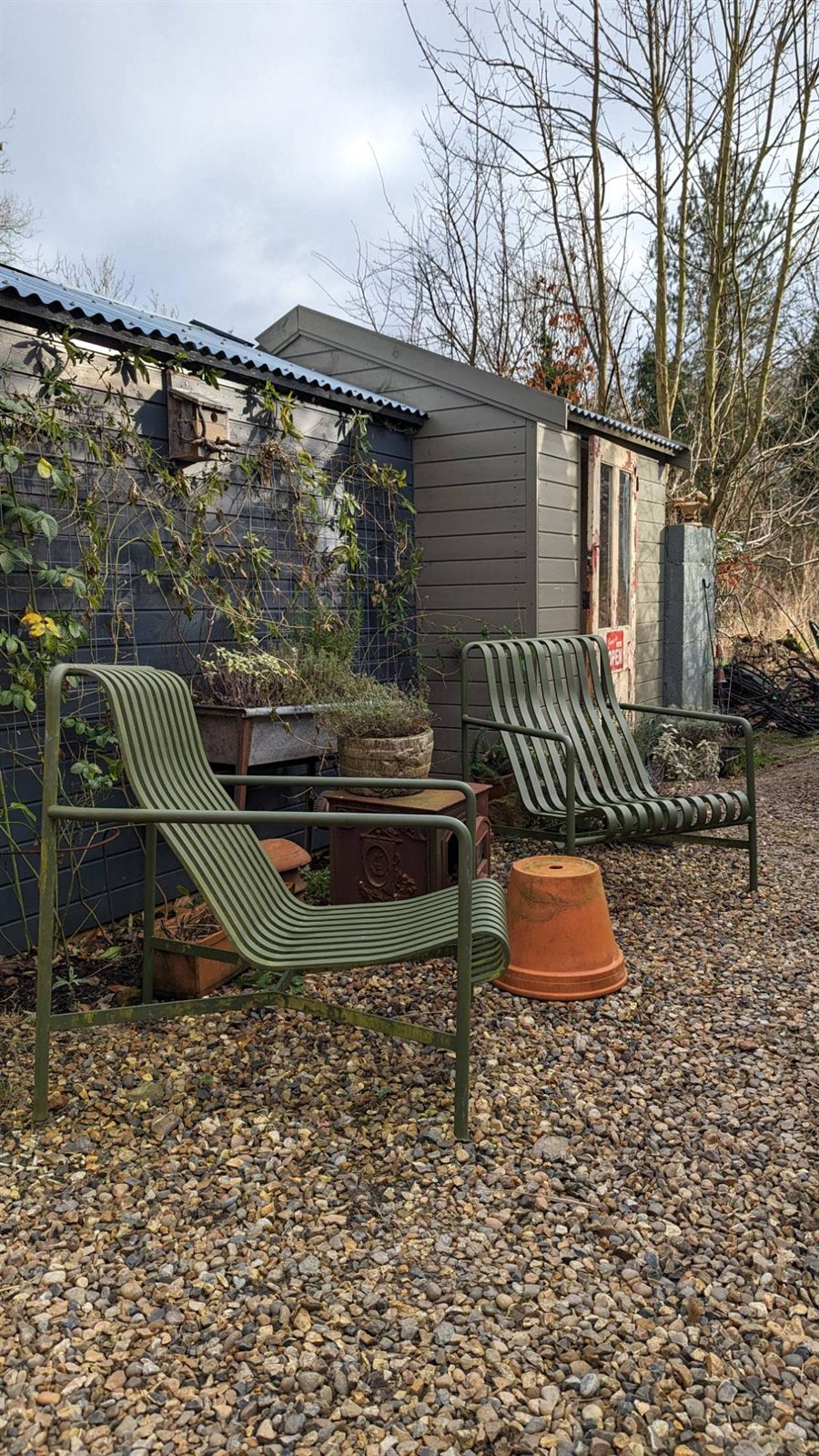 Two olive green slatted lounge chairs on gravel in front of the shed shop, with its grey-green boarded walls and weathered cream and red door behind