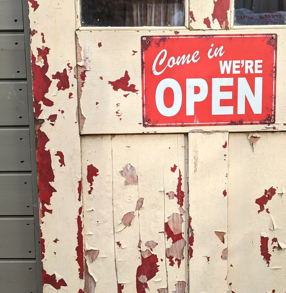 Weathered cream and red painted door of the shed shop with a red 'Come in, we're open' sign