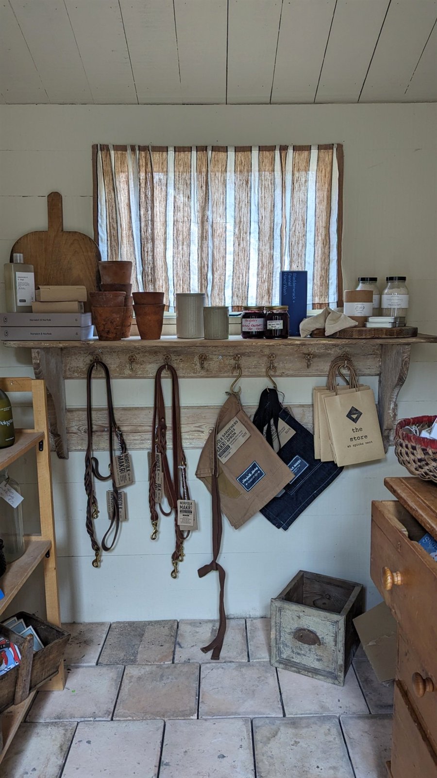 Shed shop wall with leather dog leads, canvas aprons and The Store paper bags hanging from hooks above a shelf, on a floor of terracotta pamment tiles