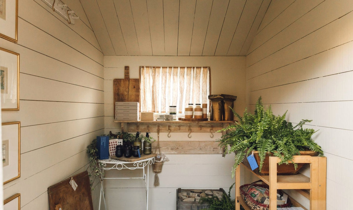 Inside the shed shop: white boarded walls and ceiling, a shelf of bath products and candles under a striped curtain, antique bread boards, a fern and a pine shelving unit